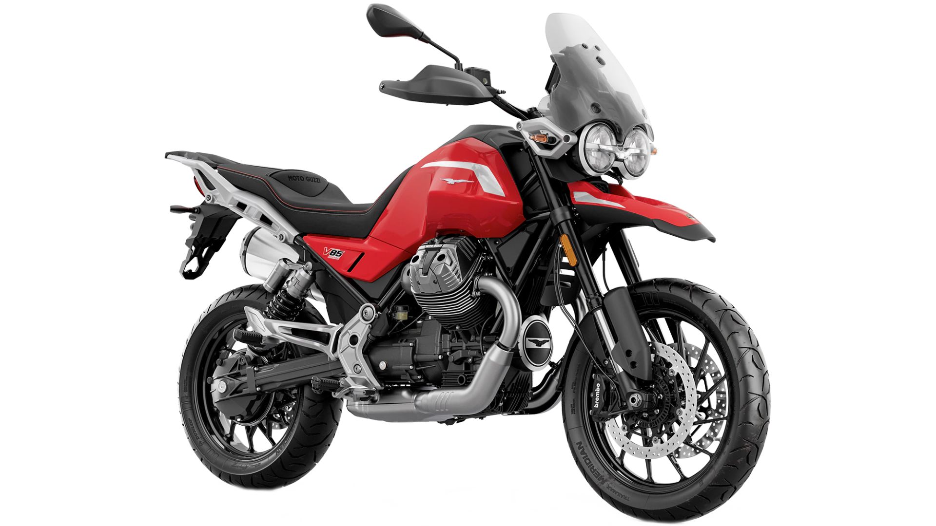 Moto Guzzi V85 TT parkiran ob marini ob sončnem zahodu, s plovili v ozadju in svetlobo, ki se odbija od vode.