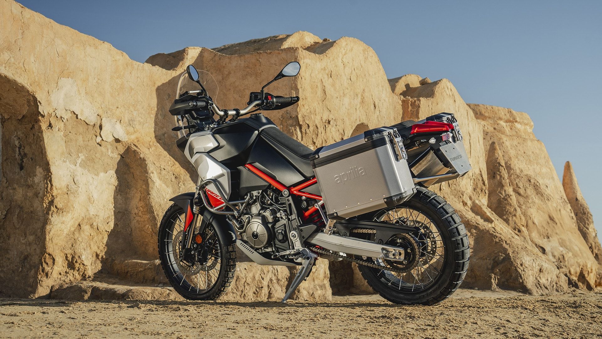 Podroben prikaz sprednjega dela motocikla Aprilia Tuareg 660 z LED žarometi, vetrobranom in krmilom, v ozadju pa razpršeno blato in prah.