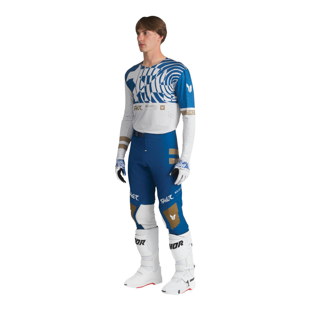Model nosi plave motocross hlače Thor Sportmode Mind Control, prikazane s prednje strane, sa zlatnim i bijelim detaljima te bijelim motocross čizmama.