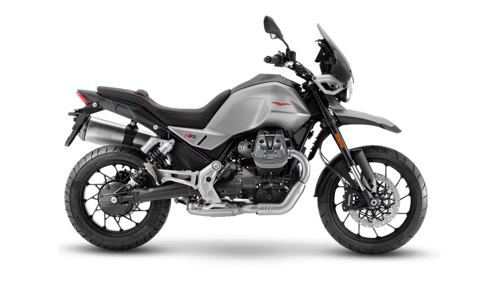 Stranski profil motocikla Moto Guzzi V85 TT v sivi barvi z rdečim logotipom, dvema izpušnima cevema, prednjim in zadnjim diskovnim zavoram ter litimi platišči, na belem ozadju.
