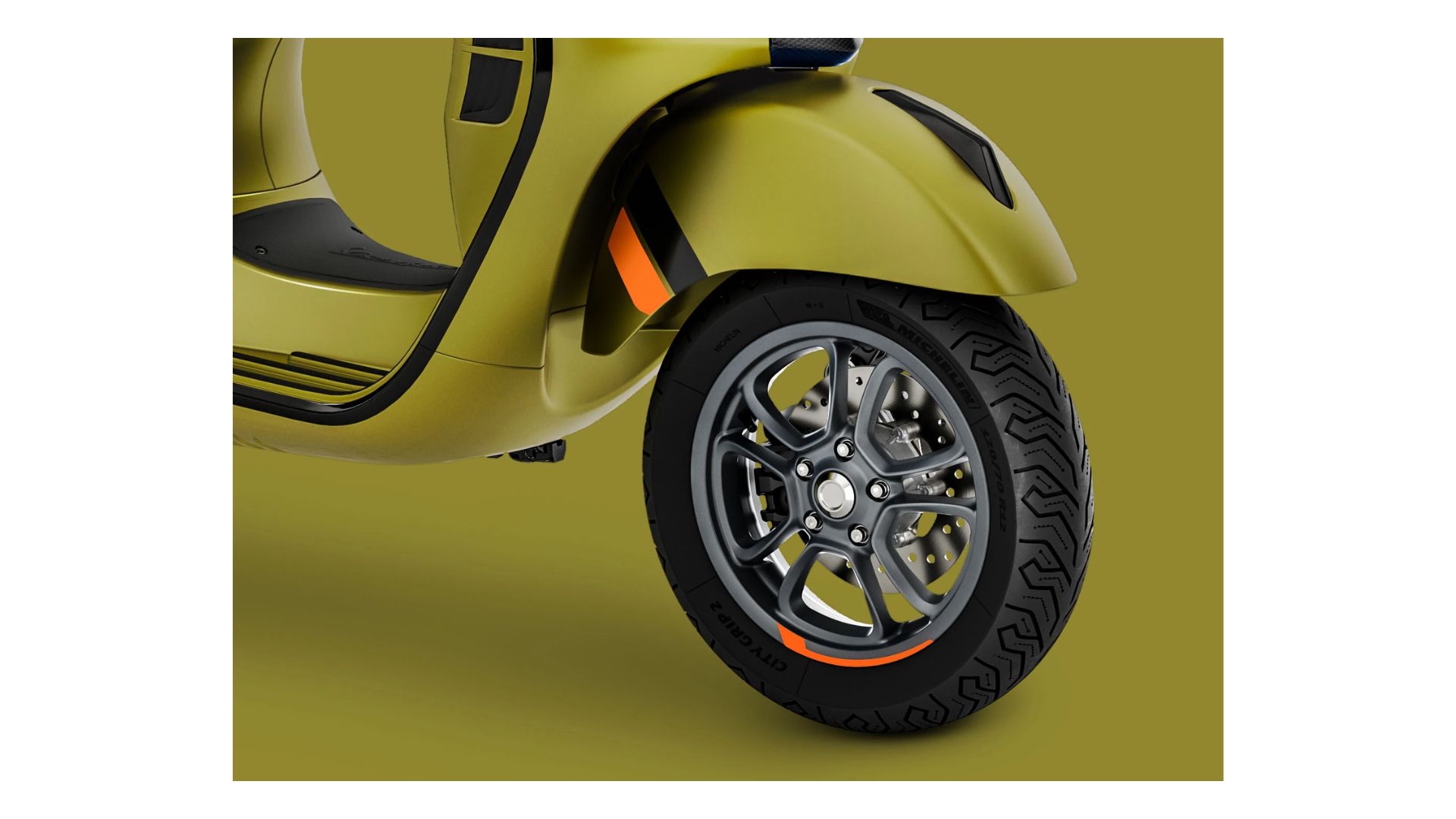 A close-up shot of the scooter's front wheel, emphasizing its alloy rim with a sleek design and Michelin tires. The prominent orange detailing on the rim stands out against the scooter's overall greenish-yellow color.